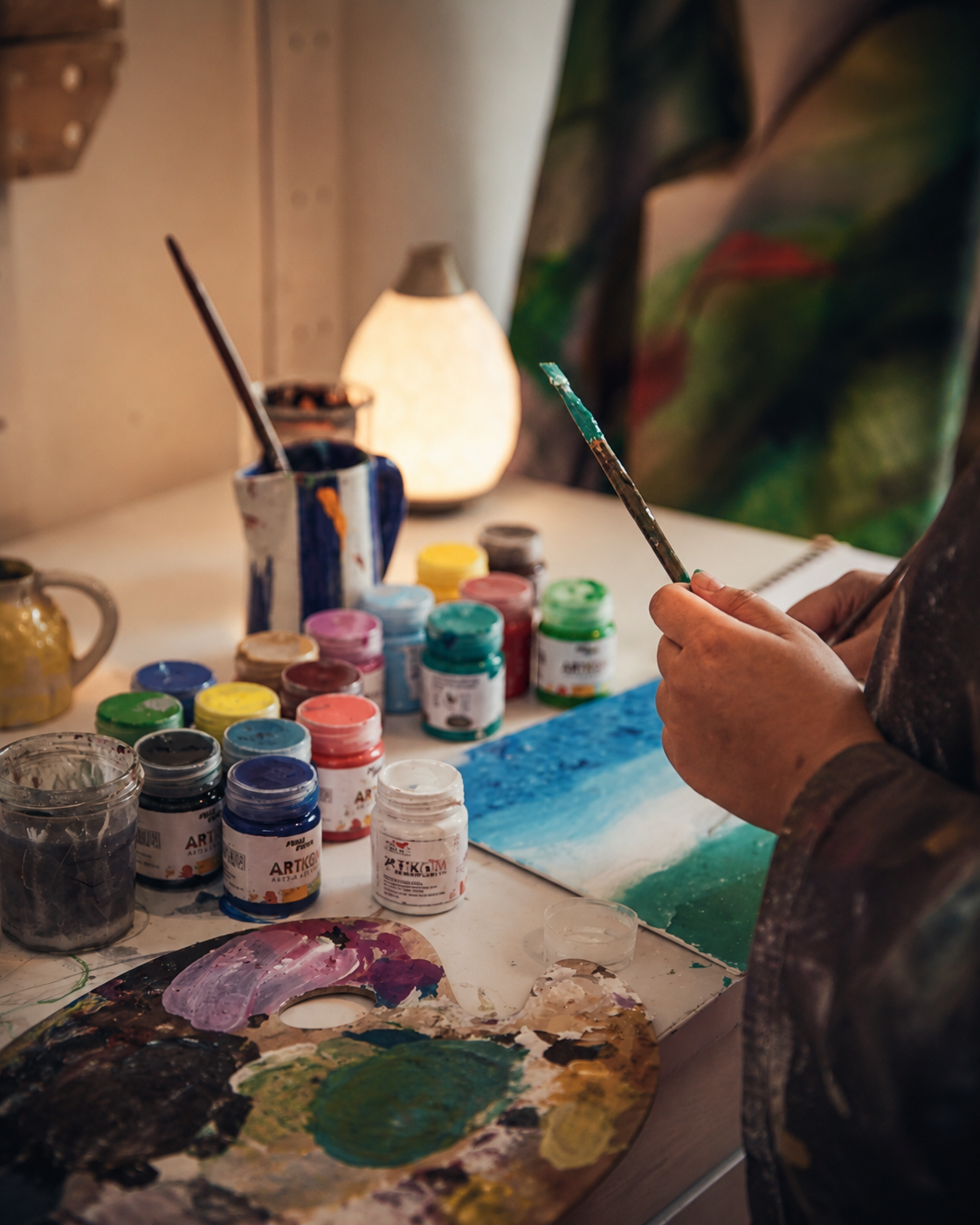 An artist's palette, paint pots and brush in warm studio light at the ARTKOM atelier.