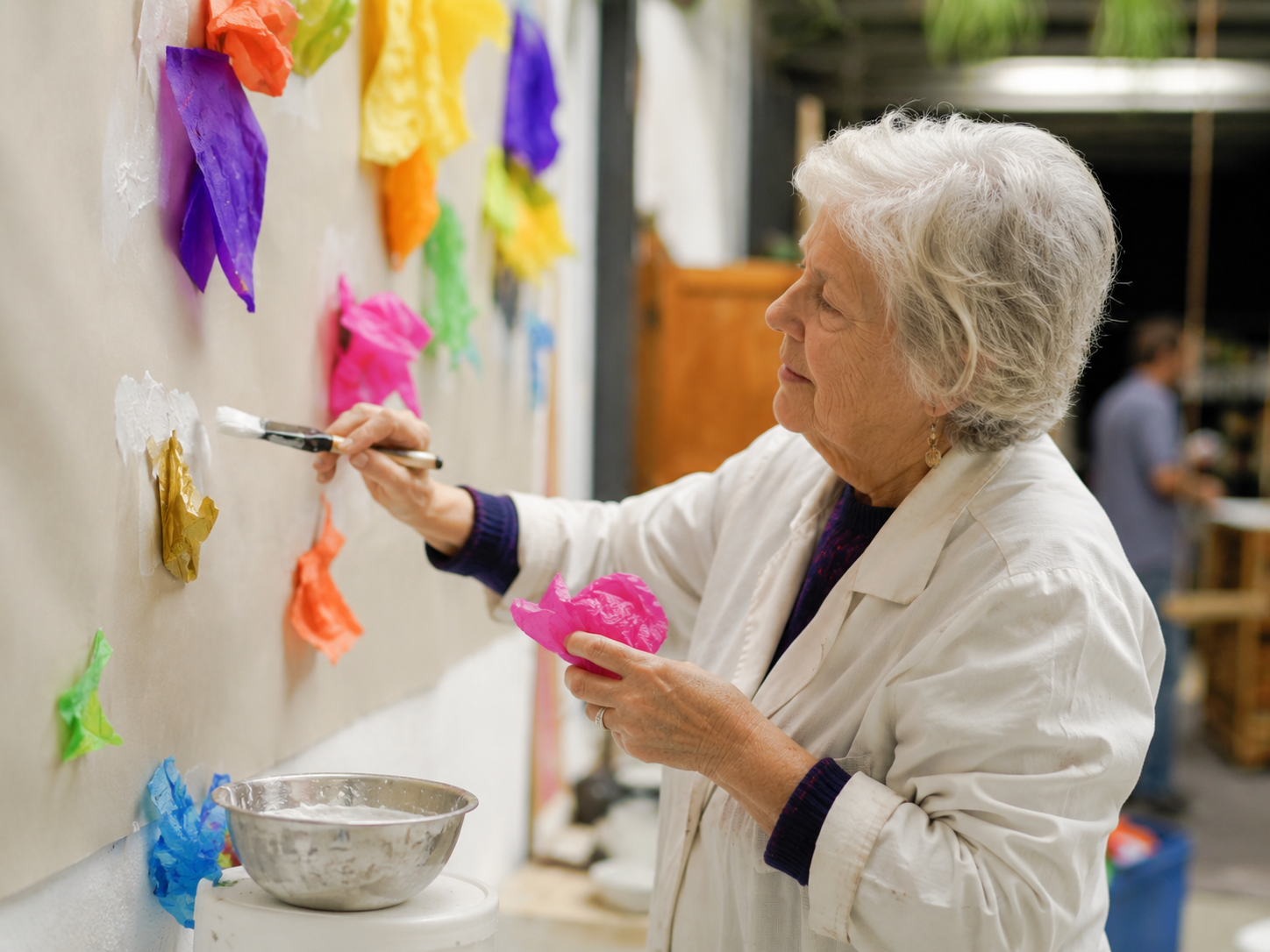 An older participant carefully brushes coloured tissue onto a wall of collective marks during an Arts & Health participatory session.