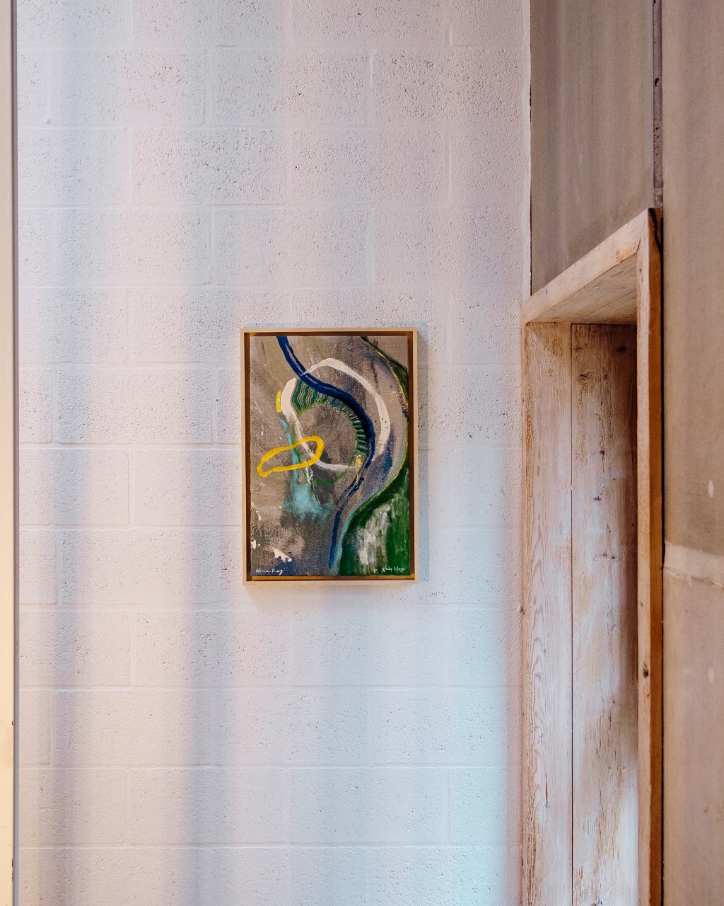 Framed painting by Nicky Myny on a white brick wall between wooden shelving and metal slats at DAFT.